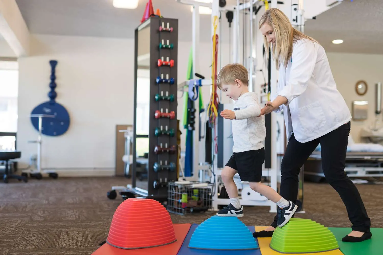 Physical therapist supporting a toddler stepping across coloured balance stones in a clinic gym