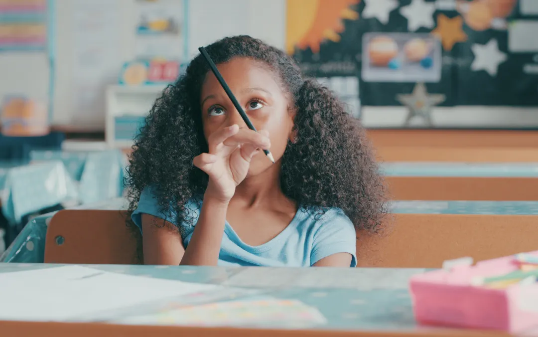Young girl at her classroom desk holding a pencil and staring off, distracted from her work