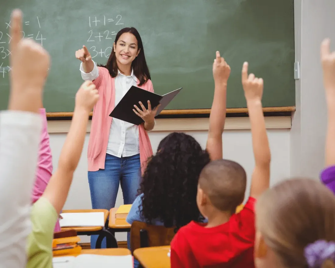 A teacher at the blackboard with a class of children raising their hands