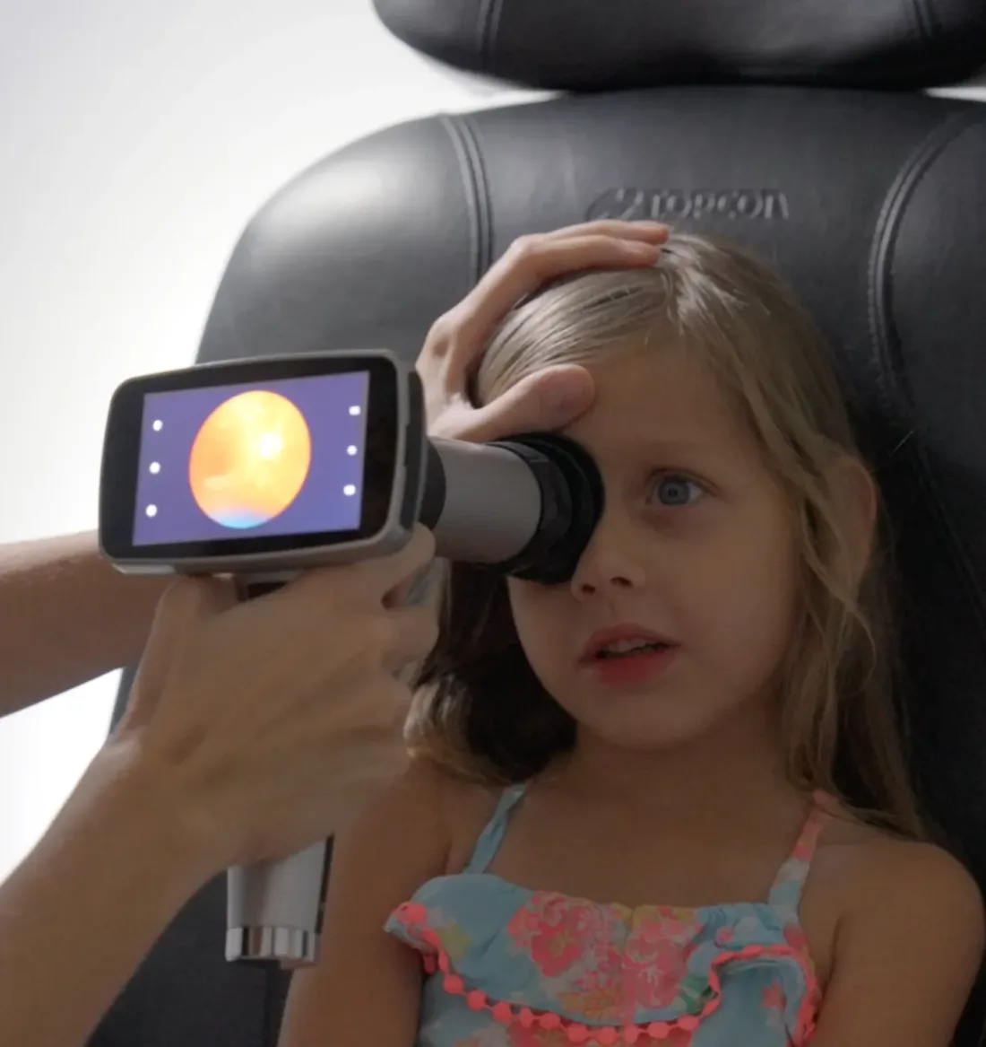 A handheld retinal camera held up to a child's eye, the retinal image showing on its screen