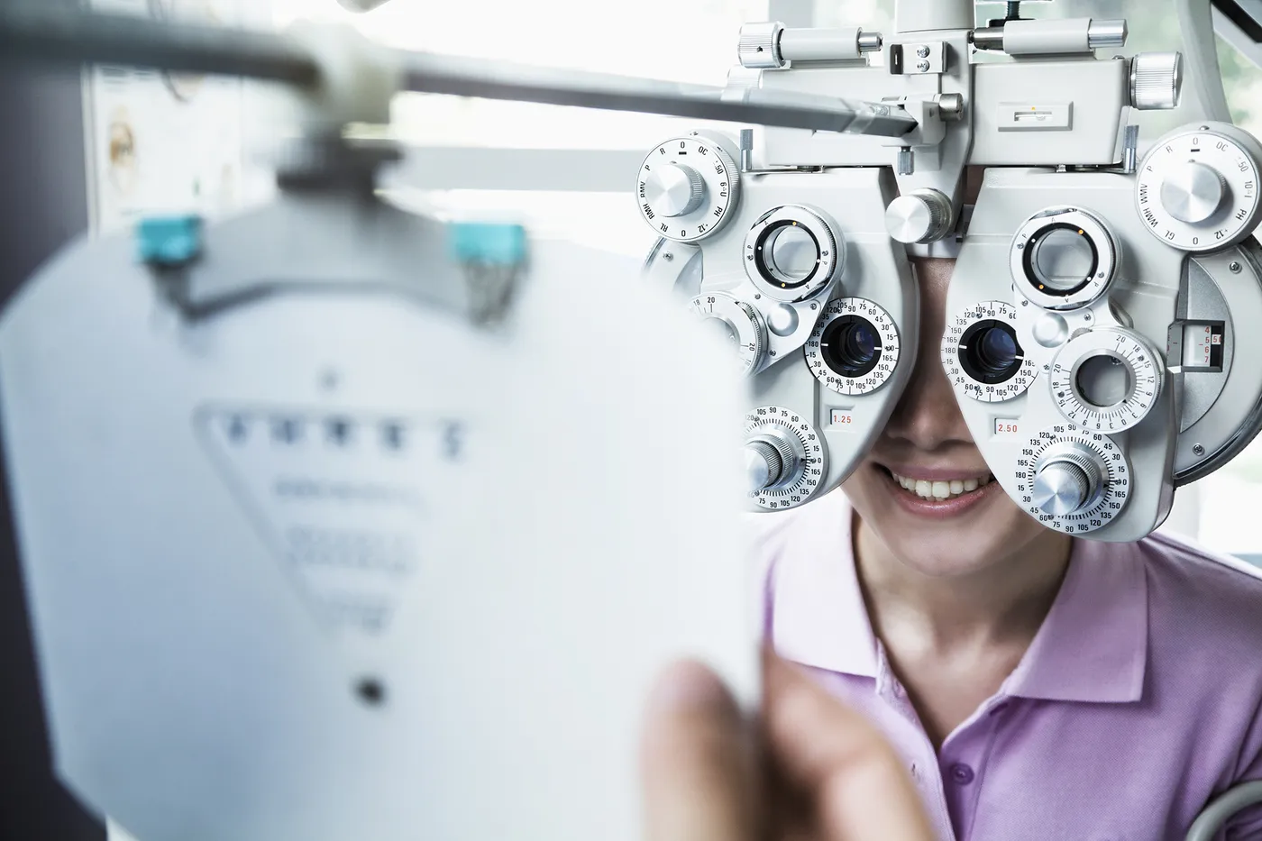 A patient looks through a phoropter during a vision evaluation, with a blurred acuity chart in the foreground