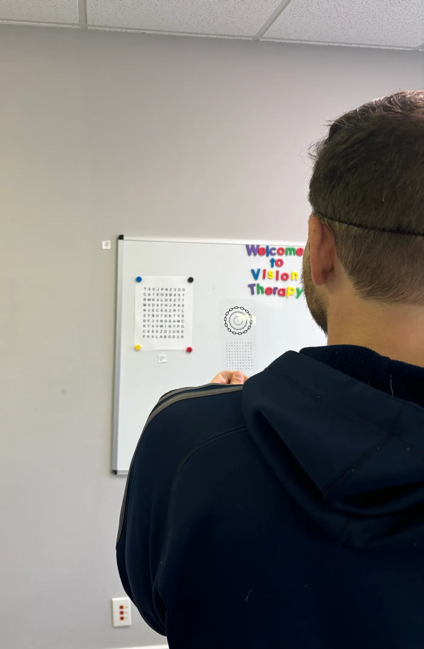A vision therapy patient reaching toward a wall-mounted board holding a large Hart chart of letters and numbers, a smaller chart and a rotation target