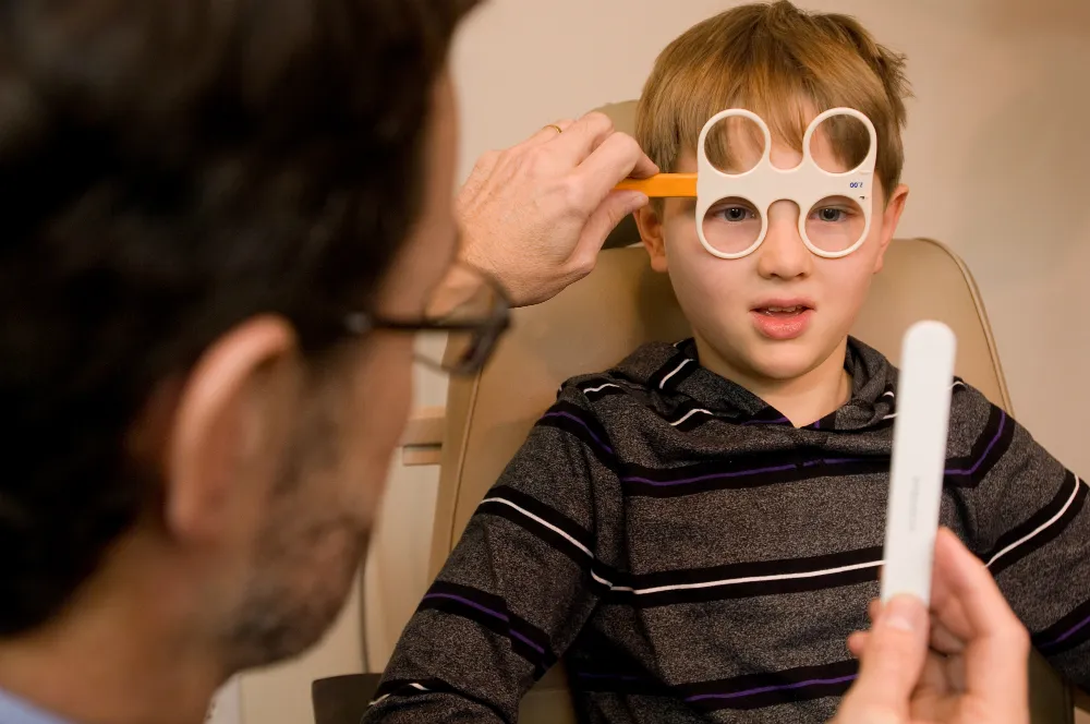 A boy wearing a loose-lens trial frame during binocular vision testing while the optometrist holds a fixation target beside his eye