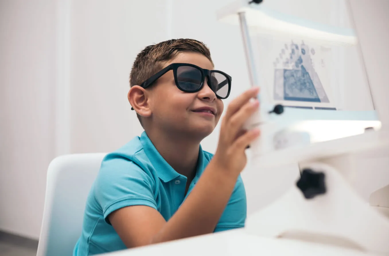 A boy in a blue shirt wearing dark therapy glasses smiles while working with a vision therapy device in a bright white therapy room.