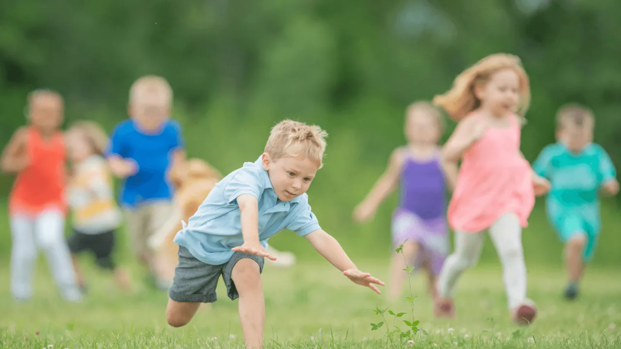 Group of children running across a grassy field in bright clothing while one boy in a blue shirt stumbles and falls forward.