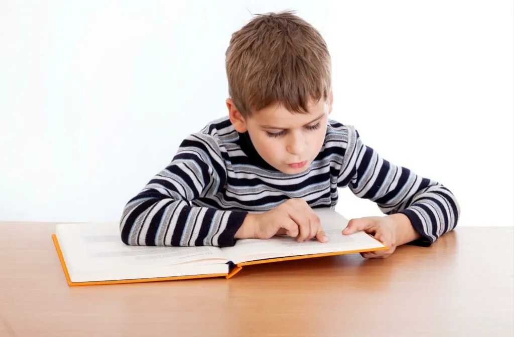 A boy in a striped sweater leans close over an open book on a table, holding his place on the line with his finger.