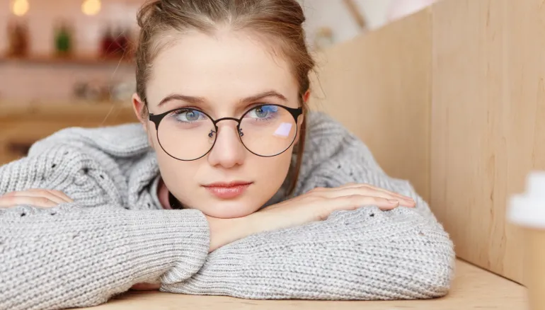 Woman wearing glasses resting her head on folded arms after prolonged near work, in a grey sweater against a warm blurred background.