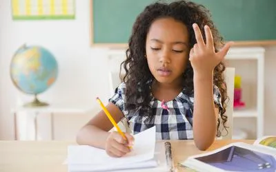 A girl at a classroom desk pauses her pencil to count on her fingers while working through a math problem