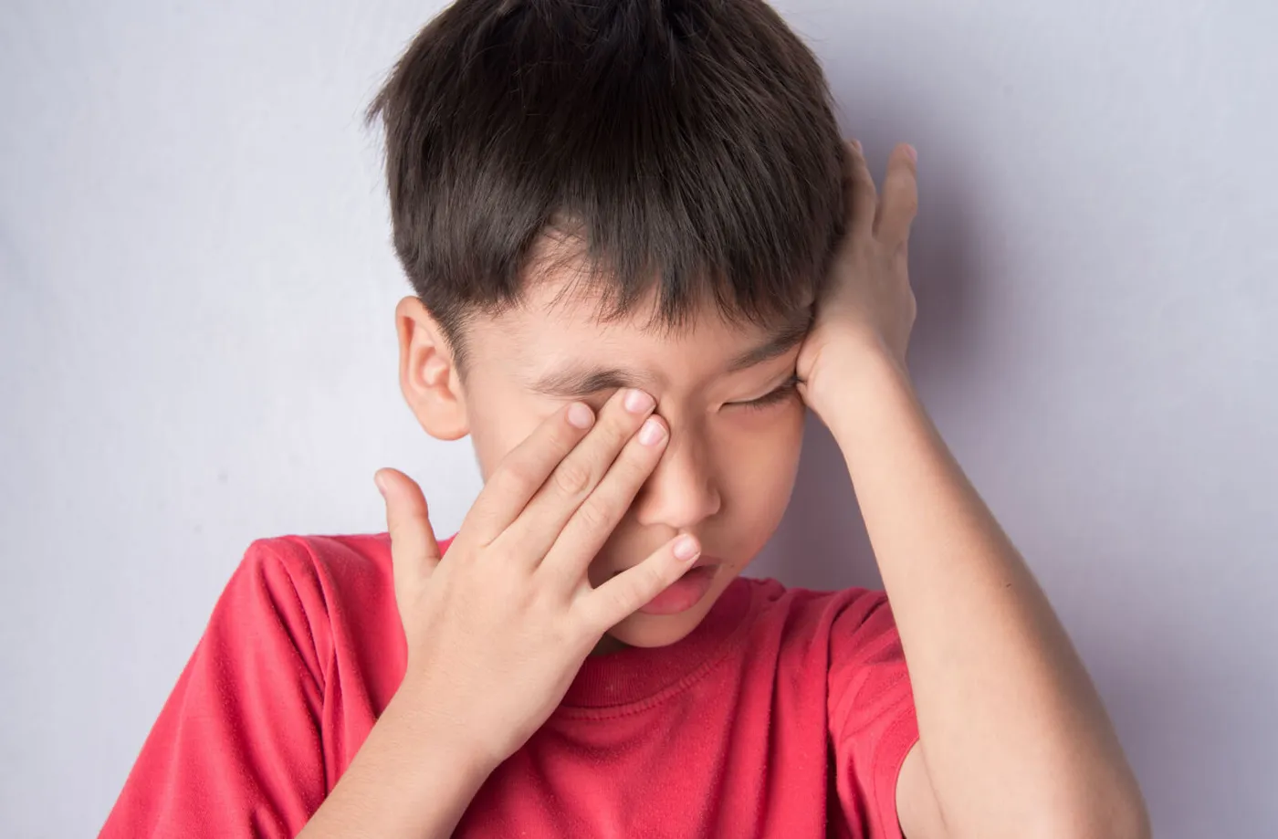 Child in a red shirt rubbing both eyes and looking tired after near work, a common end-of-homework sign families notice at home rather than in the exam room.