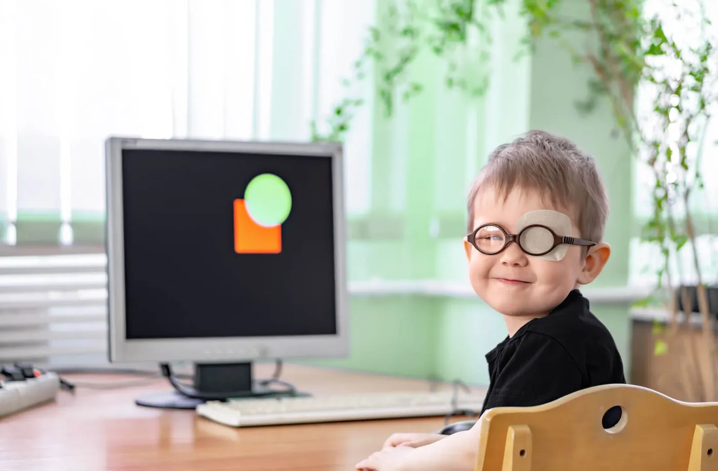 A young boy in round glasses with an adhesive patch over one lens smiles at a desk in front of a computer screen showing an orange square and a green circle during amblyopia treatment