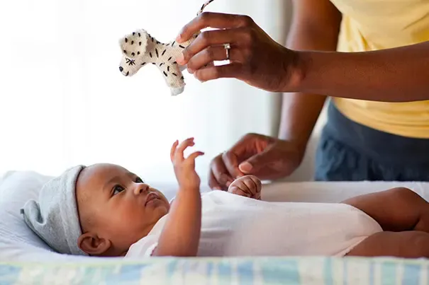 Baby lying on a changing pad, looking up and reaching one hand toward a small plush toy a parent is dangling overhead