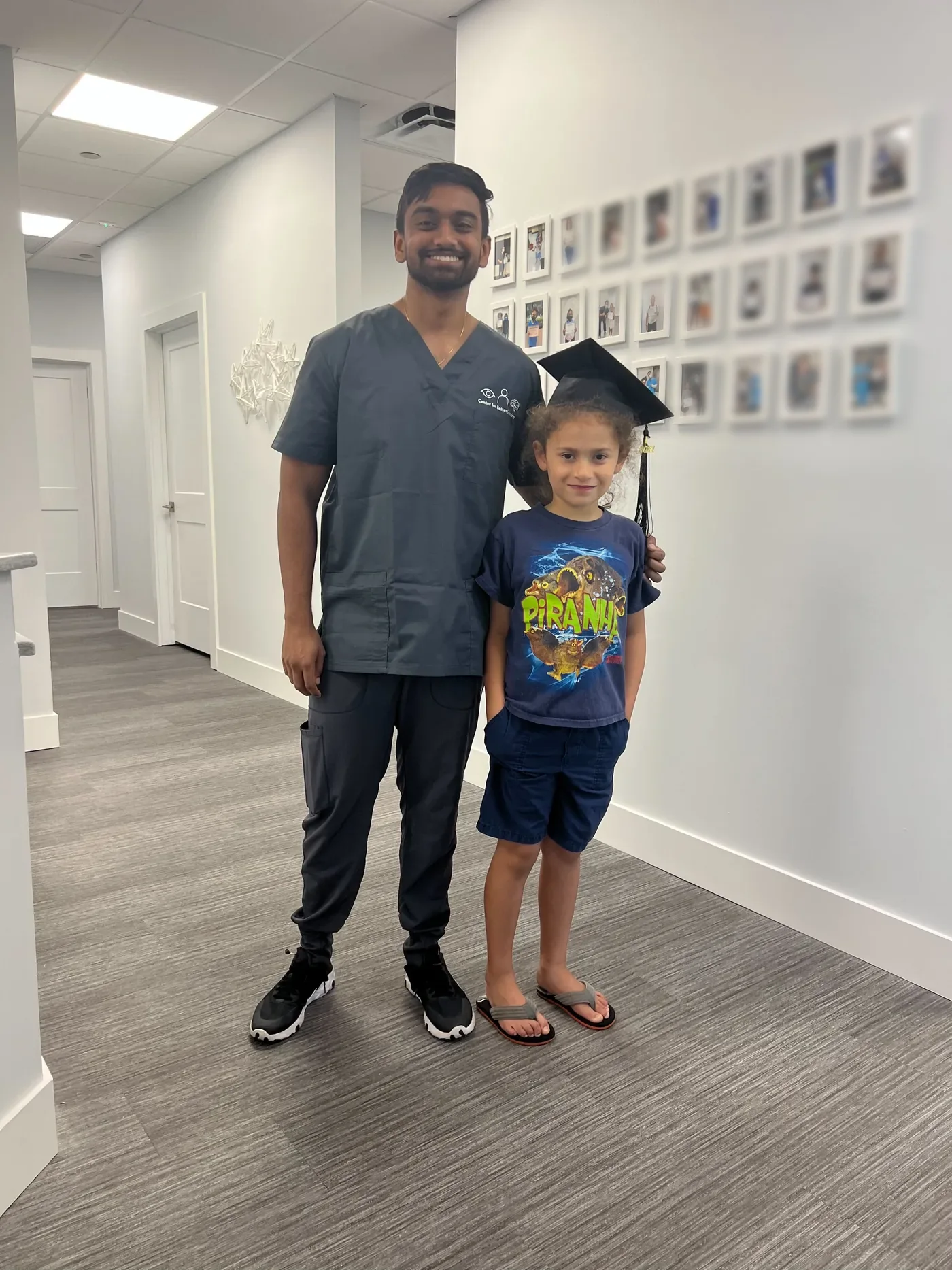A therapist stands with a young vision therapy graduate wearing a graduation cap, in front of the office wall of graduate photos