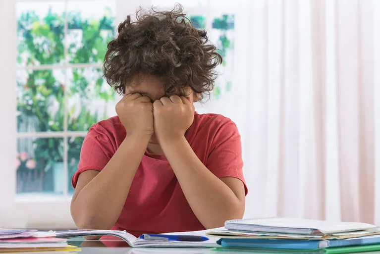 A boy at a homework table pressing his fists into his eyes over open books and papers, the kind of near-work discomfort the symptom questionnaires in these trials were built to measure