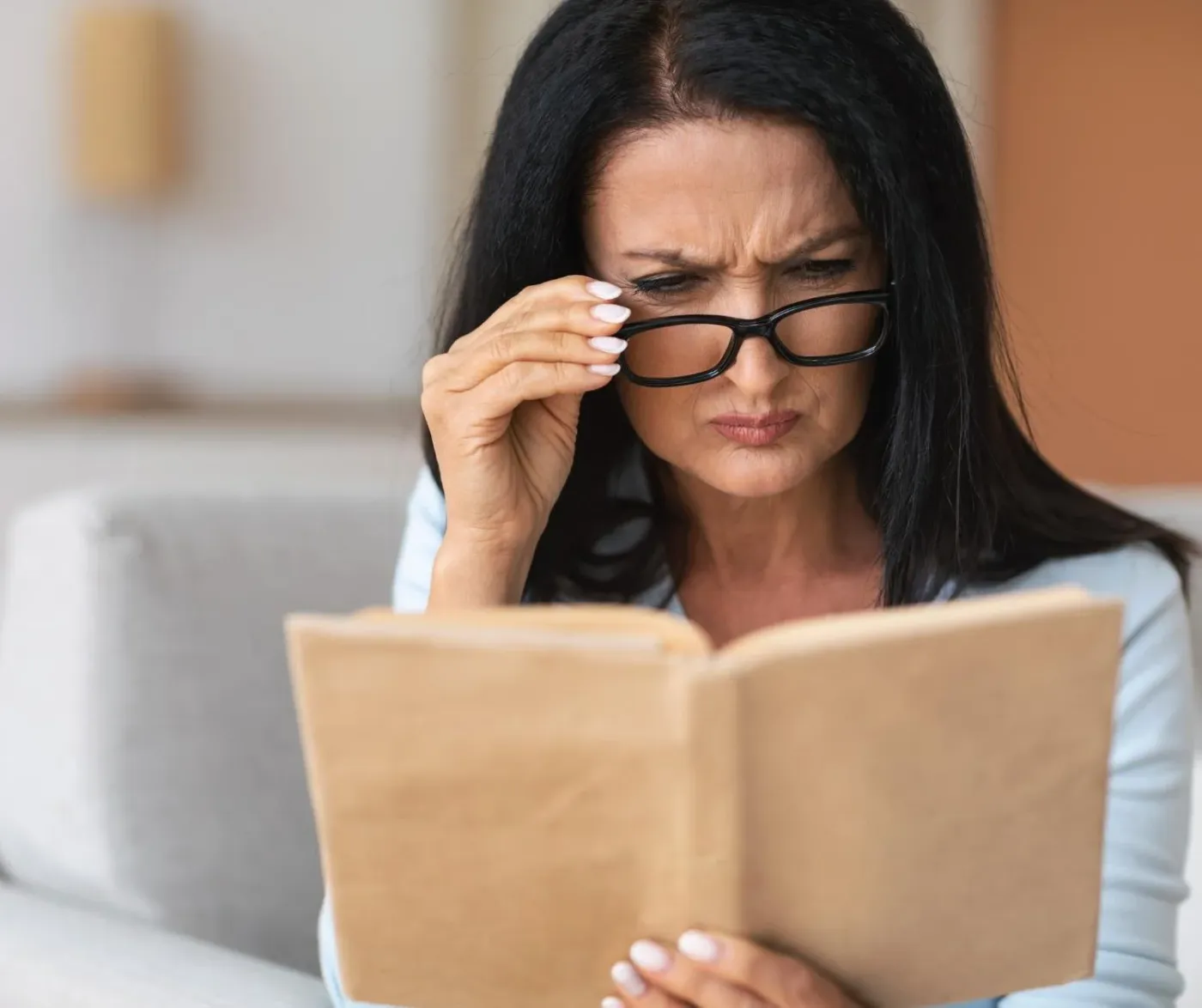 A woman in glasses frowns in concentration over a book in a softly lit room, showing that reading strain does not end in childhood.