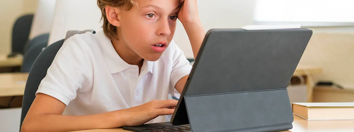 A boy in a white polo shirt leans his head on one hand at a classroom desk, looking at a tablet propped on a keyboard case, a book on the desk beside him