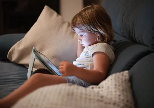 Toddler sitting on a dark sofa at night, face lit by the glow of a tablet screen