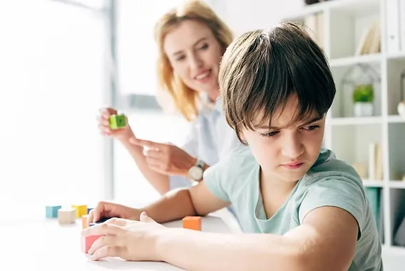 Young child in a light blue shirt looking frustrated while building with colorful blocks as an adult watches from behind