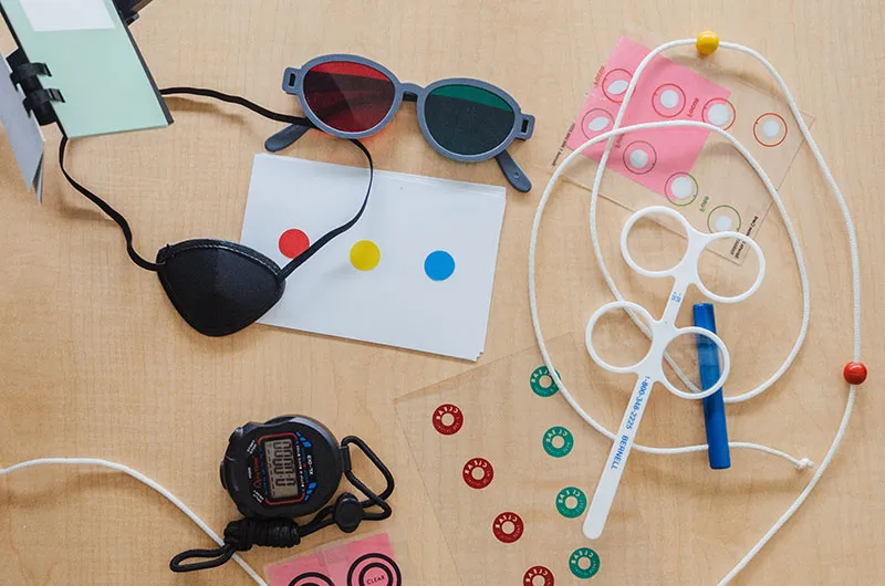 Office-based vision therapy equipment laid out on a wooden table - red-green glasses, a black eye patch, a beaded string, a loose-lens frame, colored fixation cards and a stopwatch