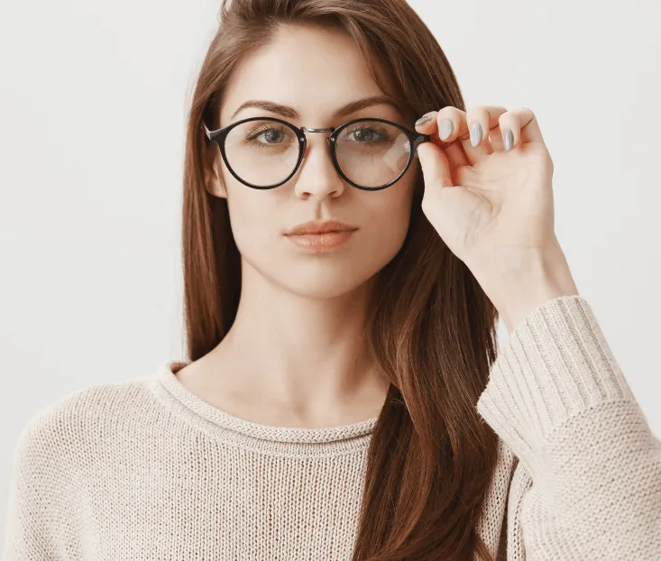 Woman with long brown hair in a beige sweater adjusting her glasses, which could carry a prism prescription, against a plain white background.