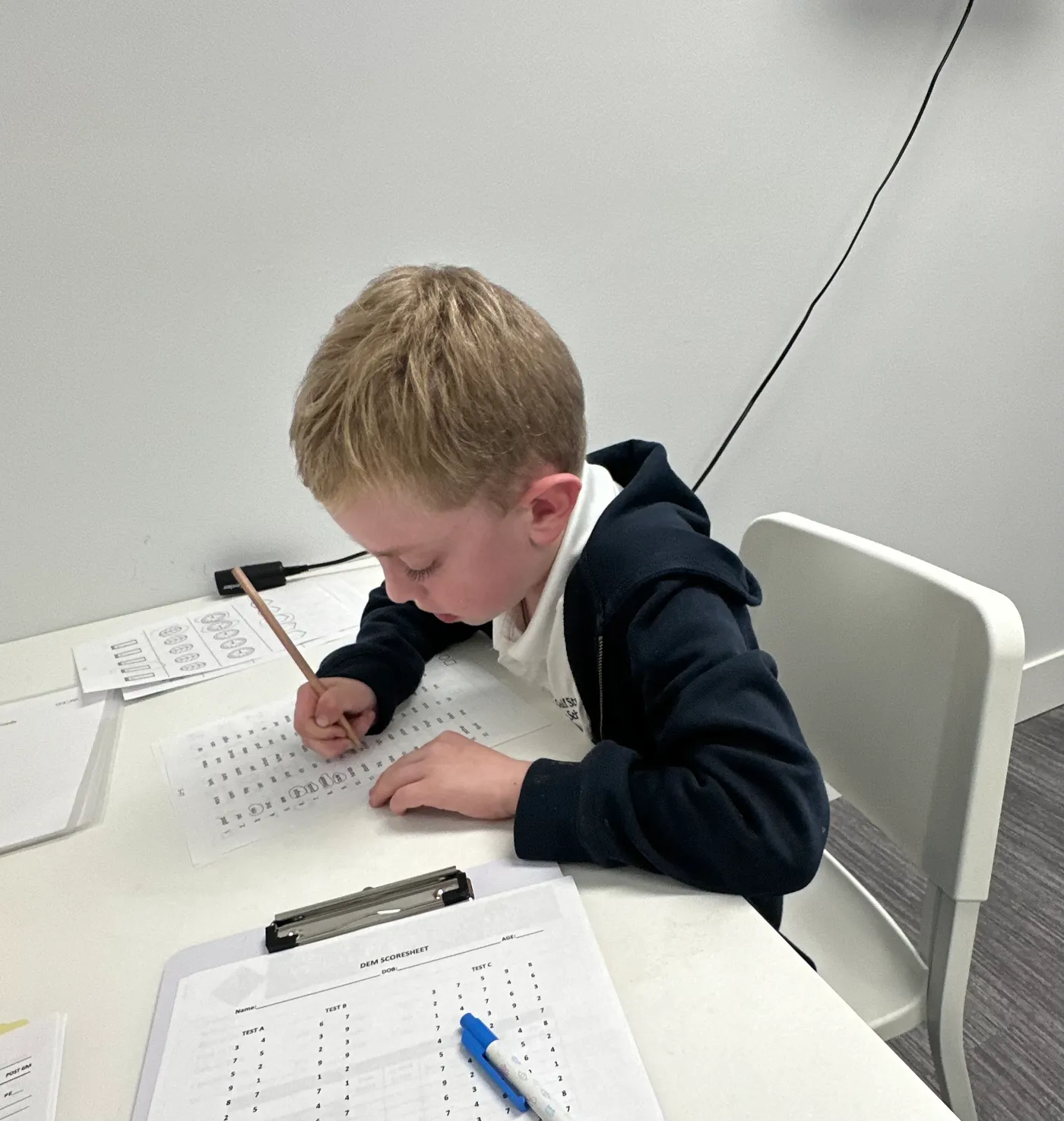 A young boy leans close to a worksheet at a white table, pencil in hand, working carefully through a page of near work.