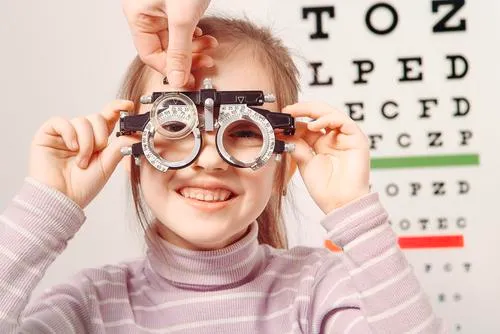 A smiling girl holding a metal trial frame to her eyes while a clinician adjusts a lens, with a Snellen letter chart on the wall behind her