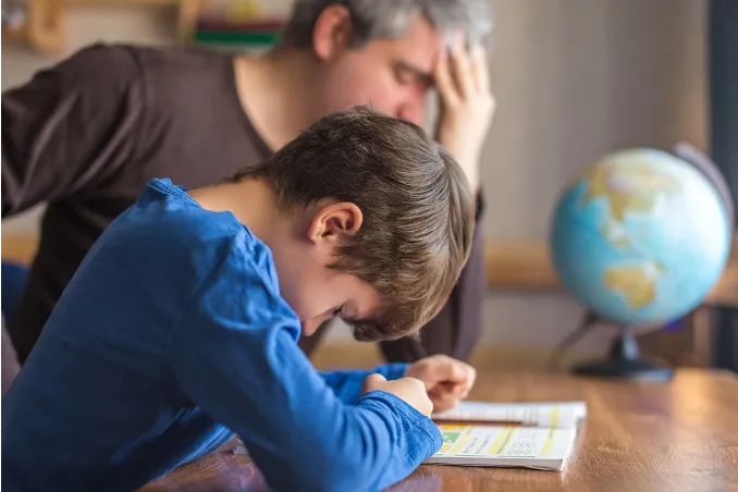 Child slumped over homework at a laptop, rubbing one eye and looking exhausted