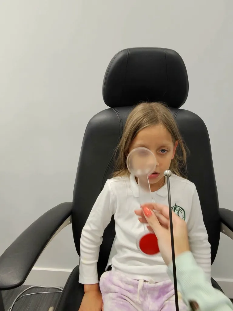 Young girl in the exam chair following a fixation target while a clinician holds a lens and a red filter in front of one eye