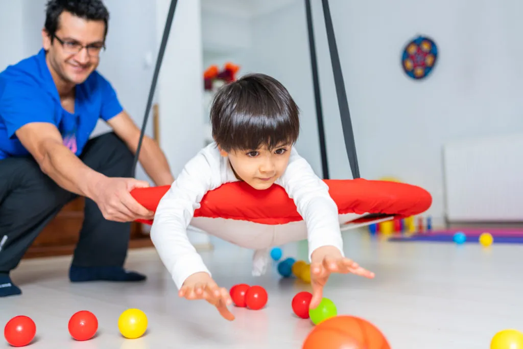 A child sits on a red platform swing in a bright therapy gym, reaching for colored balls on the floor while an adult therapist in a blue shirt steadies the swing.