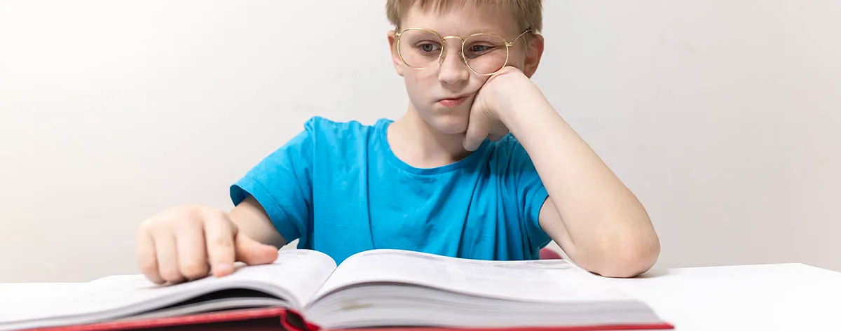 A boy in glasses slumped over an open textbook, propping his head on one hand and using a finger to keep his place on the page